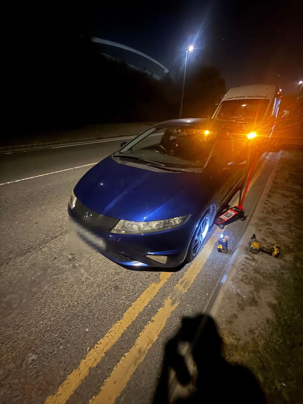 A blue Honda Civic on a jack at the roadside at night, lit by the amber beacon of a JL Mobile Tyres van