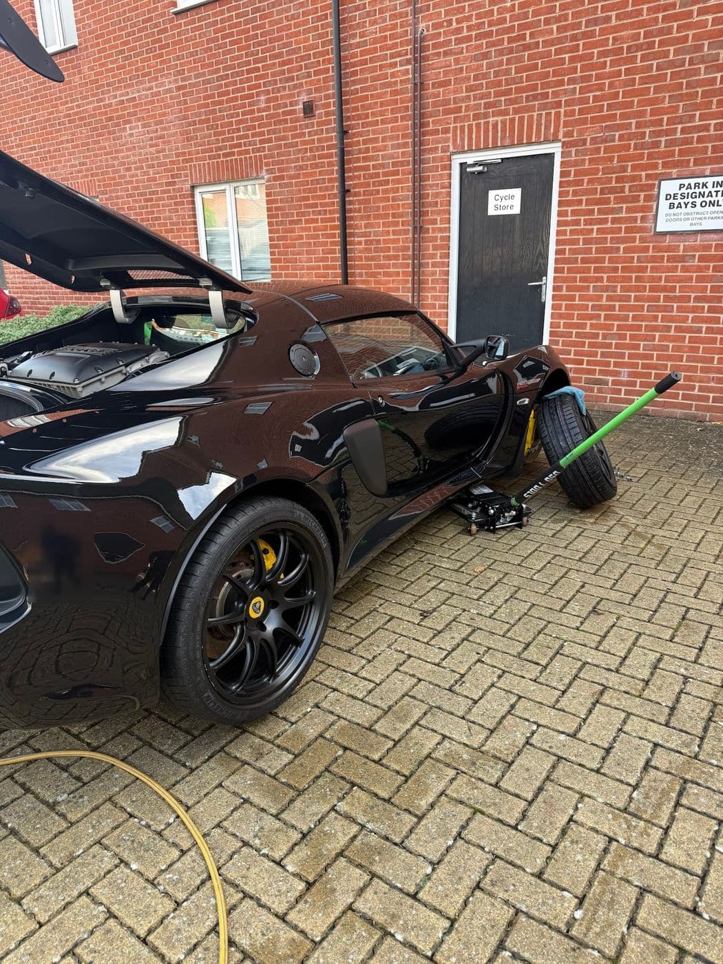 A black Lotus with its engine cover up and a wheel off, a trolley jack beside it on block paving