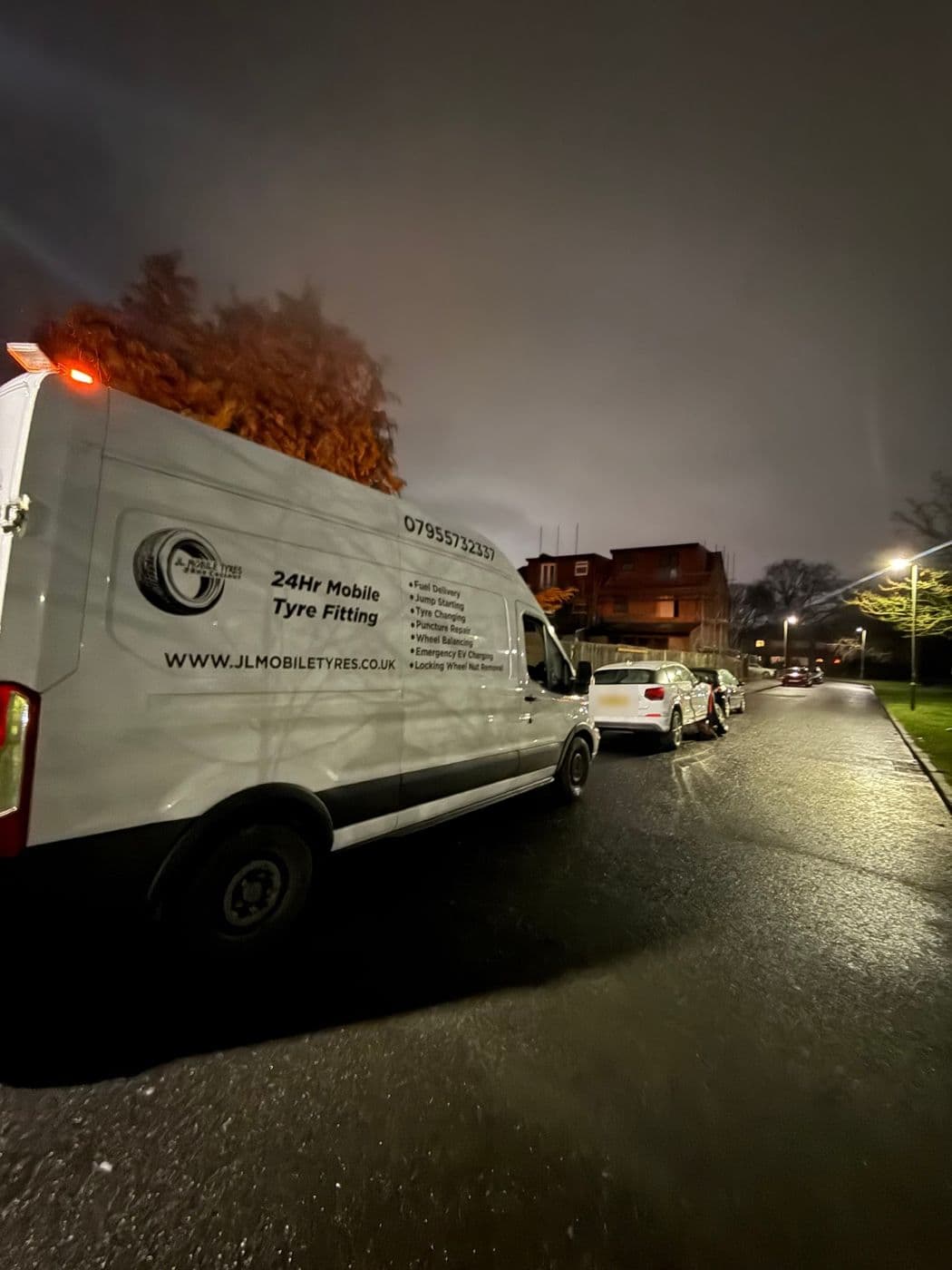 A JL Mobile Tyres van parked on a wet residential street at night, beacon on, with a white Audi further along the kerb