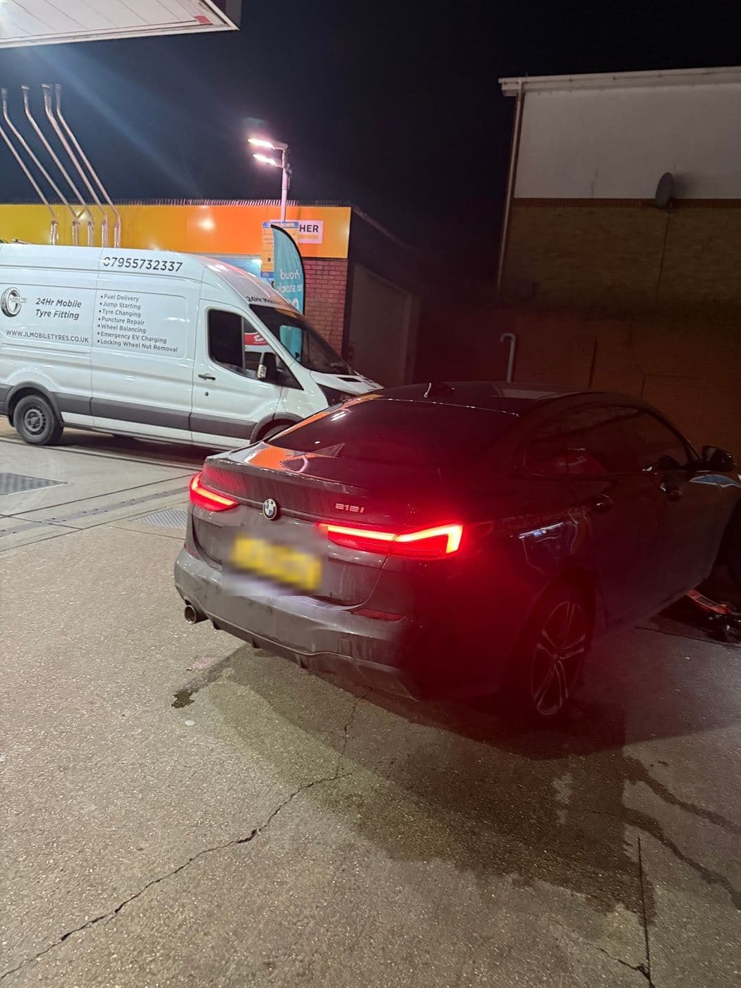 A grey BMW with its rear lights on at a petrol station forecourt at night, a JL Mobile Tyres van parked beside it