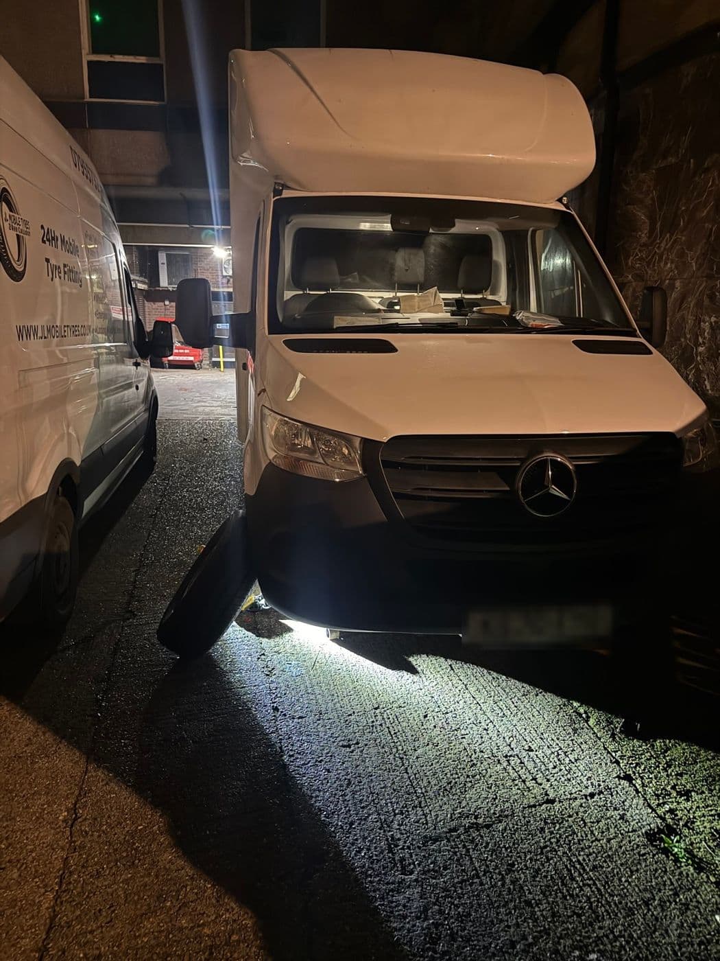 A white Mercedes Sprinter box van parked at night in a town centre service road, a JL Mobile Tyres van beside it