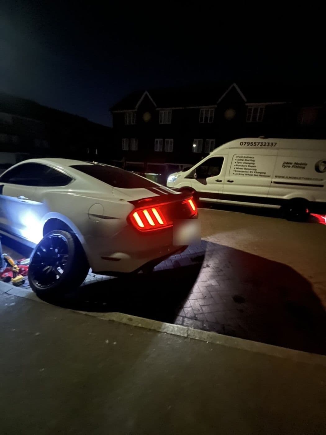A white Ford Mustang at night with its rear lights on, a JL Mobile Tyres van parked behind