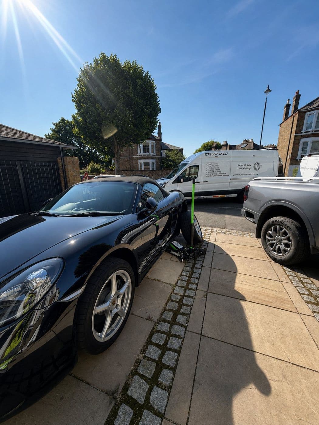 A black Porsche raised on a jack on a driveway, with a JL Mobile Tyres van in the background